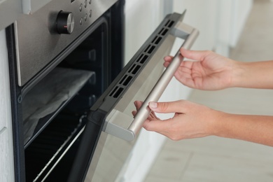 Photo of Young woman opening electric oven in kitchen, closeup