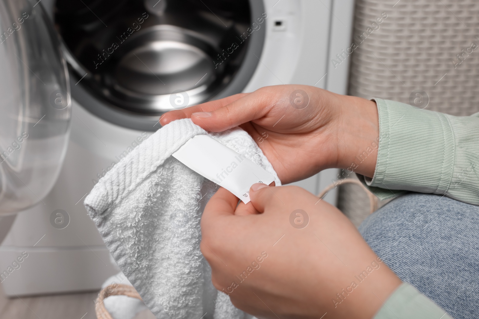 Photo of Woman reading clothing label with care symbols and material content on white towel near washing machine, closeup