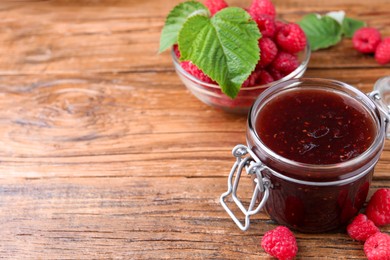 Photo of Jar of delicious raspberry jam, fresh berries and green leaves on wooden table. Space for text