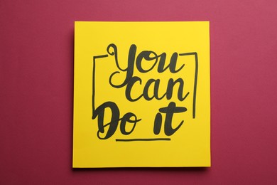 Photo of Yellow card with motivational phrase You Can Do It on burgundy background, top view