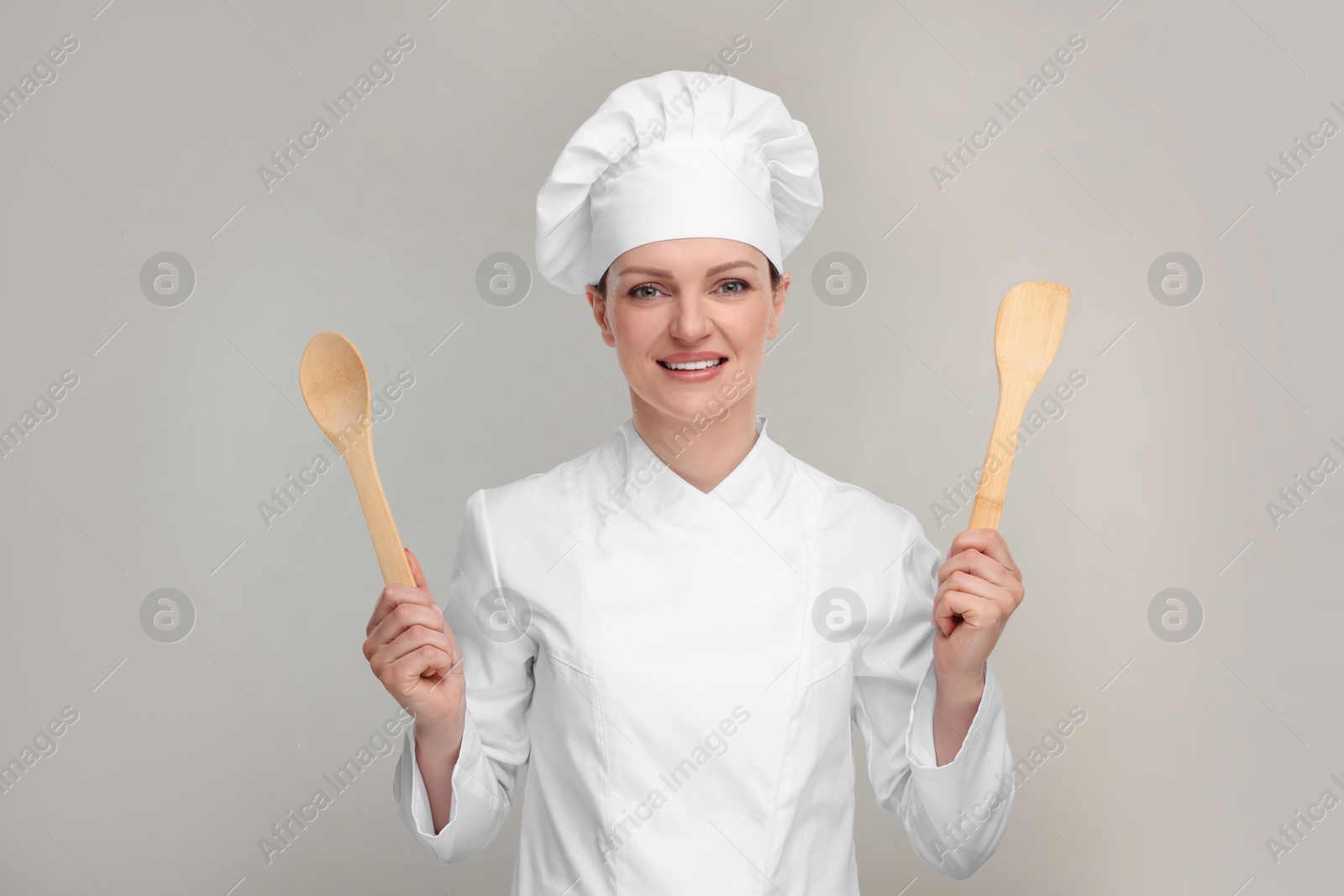 Photo of Happy woman chef in uniform holding wooden spoon and spatula on grey background