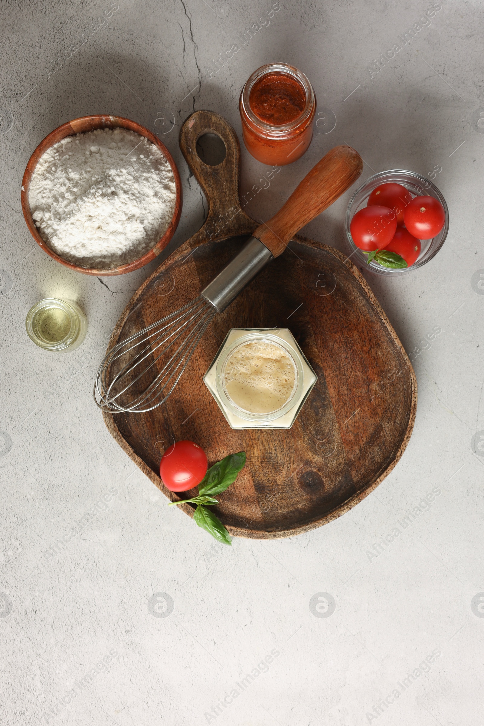 Photo of Pizza dough starter in glass jar, products and whisk on gray textured table, flat lay
