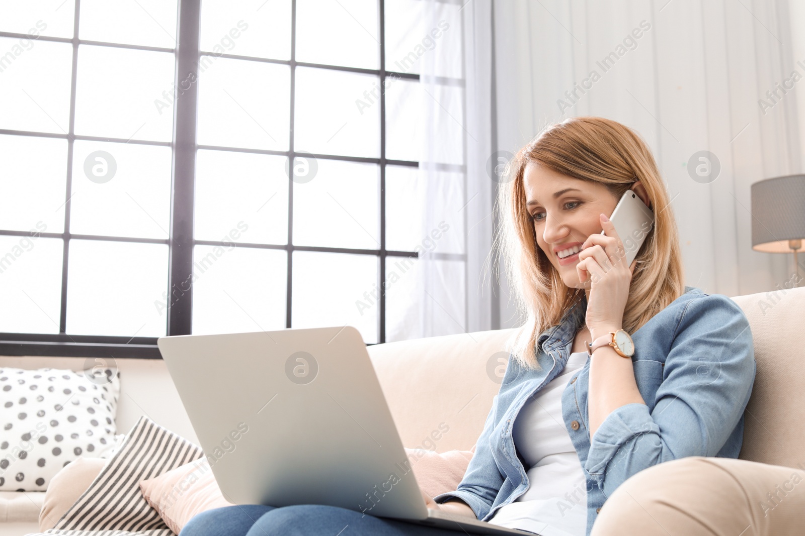 Photo of Young woman talking on phone while working with laptop on sofa in home office