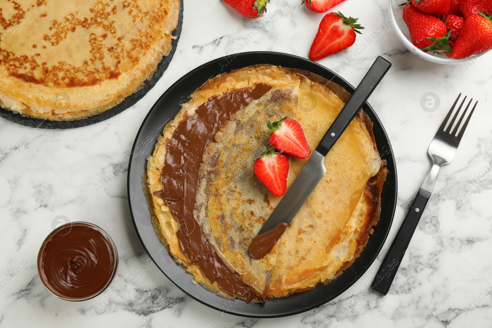 Photo of Delicious thin pancakes served with chocolate paste and strawberries on white marble table, flat lay
