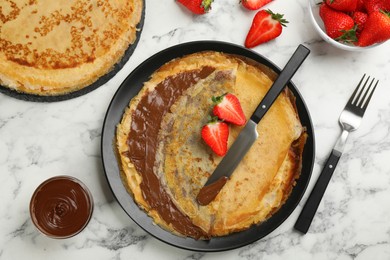 Delicious thin pancakes served with chocolate paste and strawberries on white marble table, flat lay