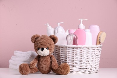 Photo of Baby cosmetic products in wicker basket, bath accessories and knitted toy bear on white table against pink background