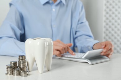 Photo of Woman using calculator at white table indoors, focus on ceramic model of tooth and coins. Expensive treatment