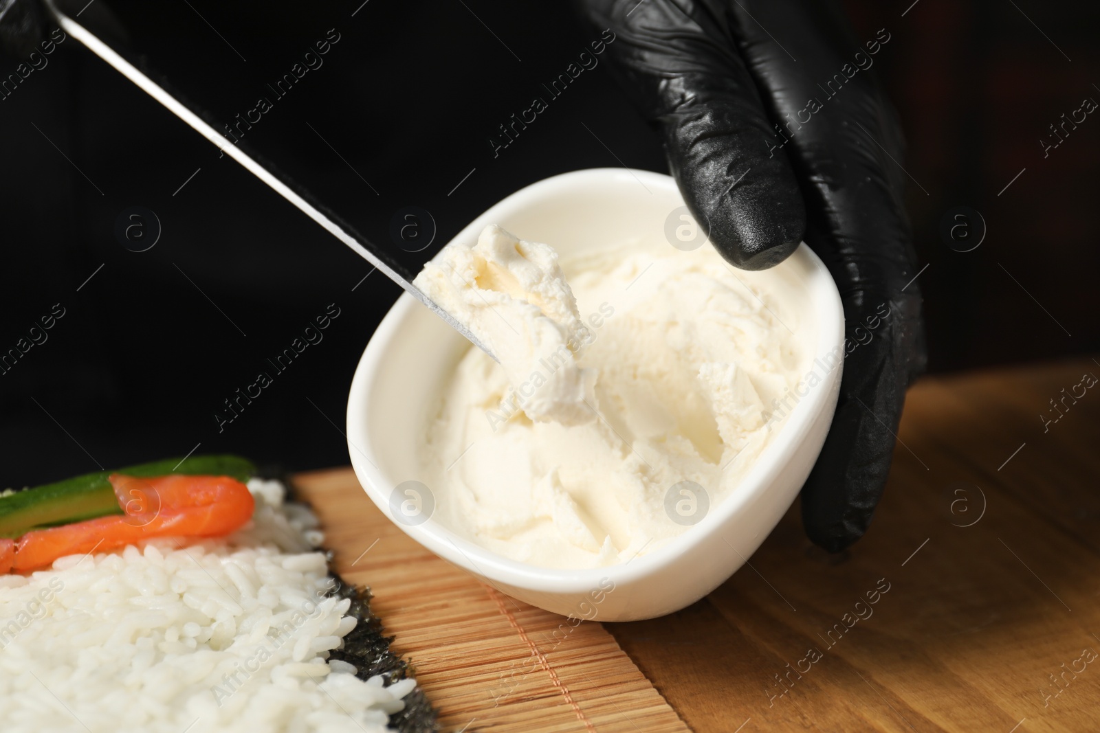 Photo of Chef in gloves putting cream cheese onto unwrapped sushi roll at wooden table, closeup