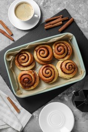 Photo of Baking dish with tasty cinnamon rolls, sticks and coffee on grey textured table, flat lay