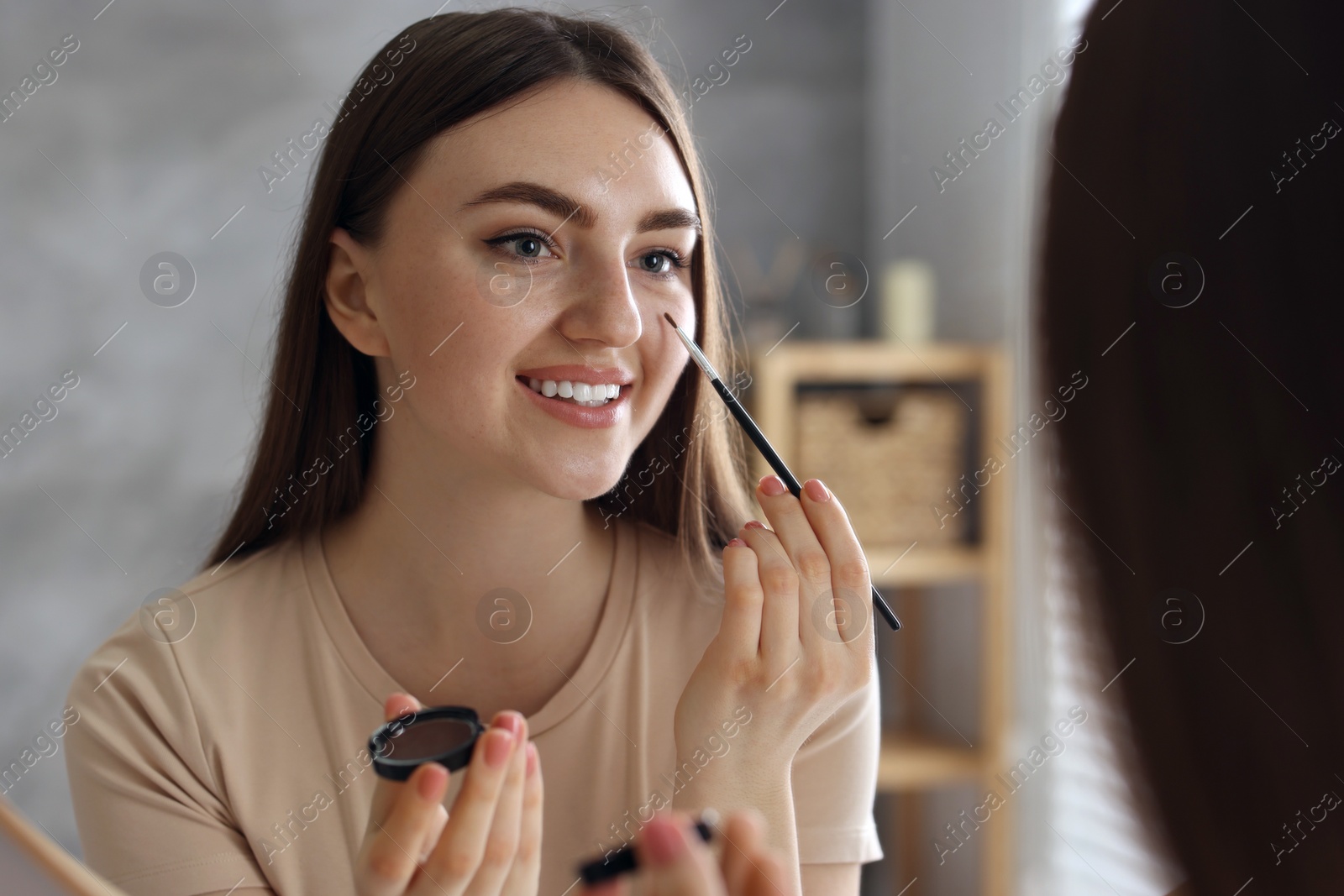 Photo of Smiling woman with freckles applying makeup near mirror indoors