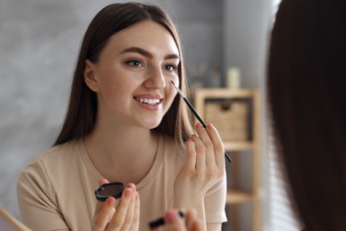 Photo of Smiling woman with freckles applying makeup near mirror indoors