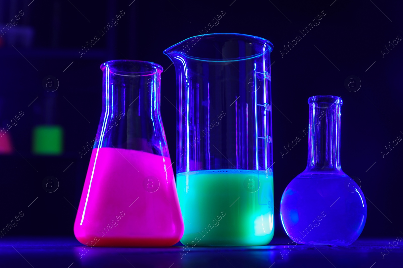 Photo of Laboratory glassware with luminous liquids on table against dark background