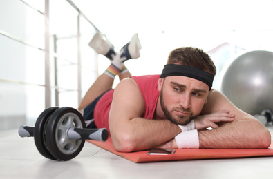 Photo of Lazy young man with abs roller lying on yoga mat indoors