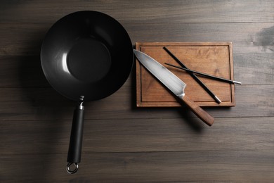 Black metal wok, knife, board and chopsticks on wooden table, top view