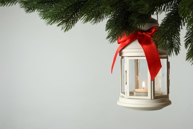 Photo of Christmas lantern with candle hanging on fir tree branch against light background. Space for text