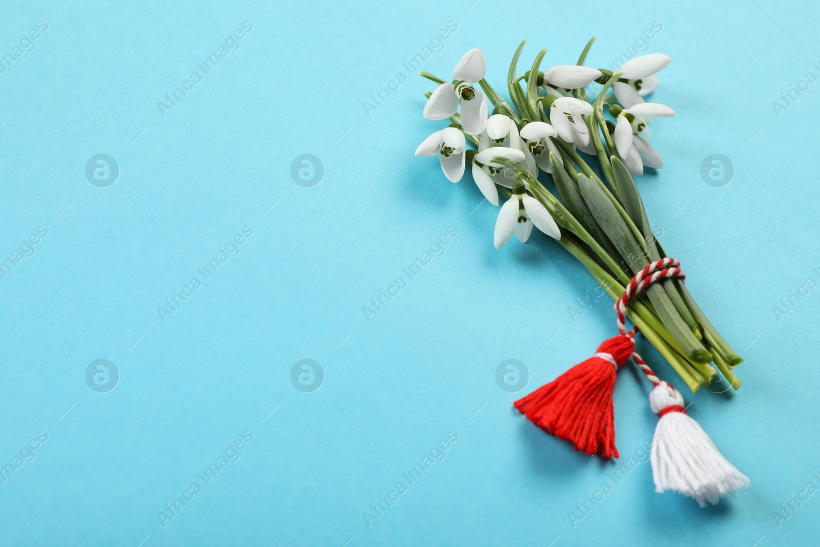 Photo of Beautiful snowdrops with traditional martisor on light blue background, flat lay and space for text. Symbol of first spring day