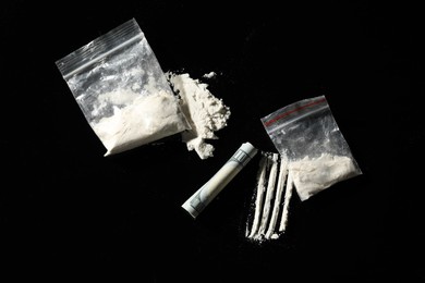 Photo of Drug addiction. Plastic bags with cocaine and rolled dollar banknote on black background, flat lay