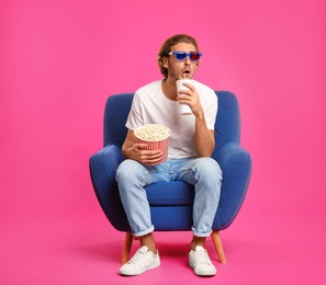 Photo of Man with 3D glasses, popcorn and beverage sitting in armchair during cinema show on color background