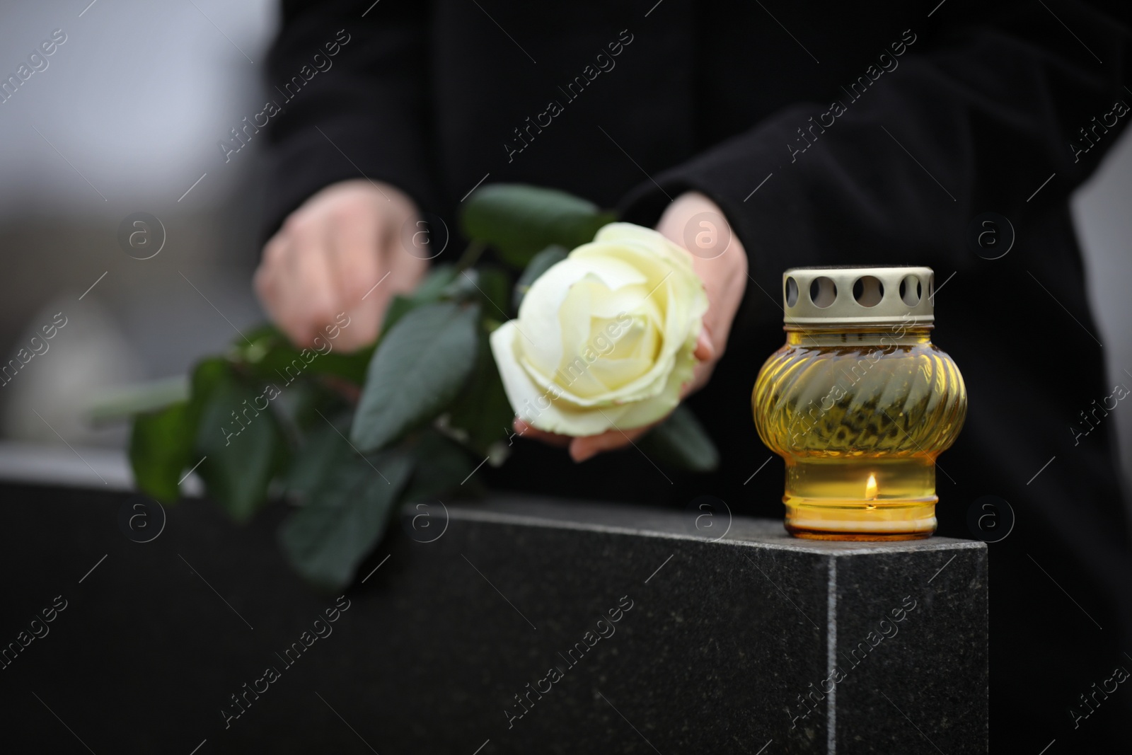 Photo of Woman holding white rose near black granite tombstone with candle outdoors, closeup. Funeral ceremony