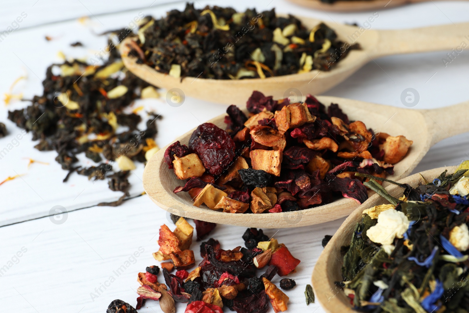 Photo of Spoons with dried herbal tea leaves and fruits on white wooden table, closeup