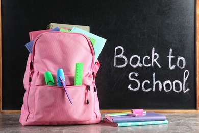 Photo of Backpack with stationery on table against blackboard with written words BACK TO SCHOOL