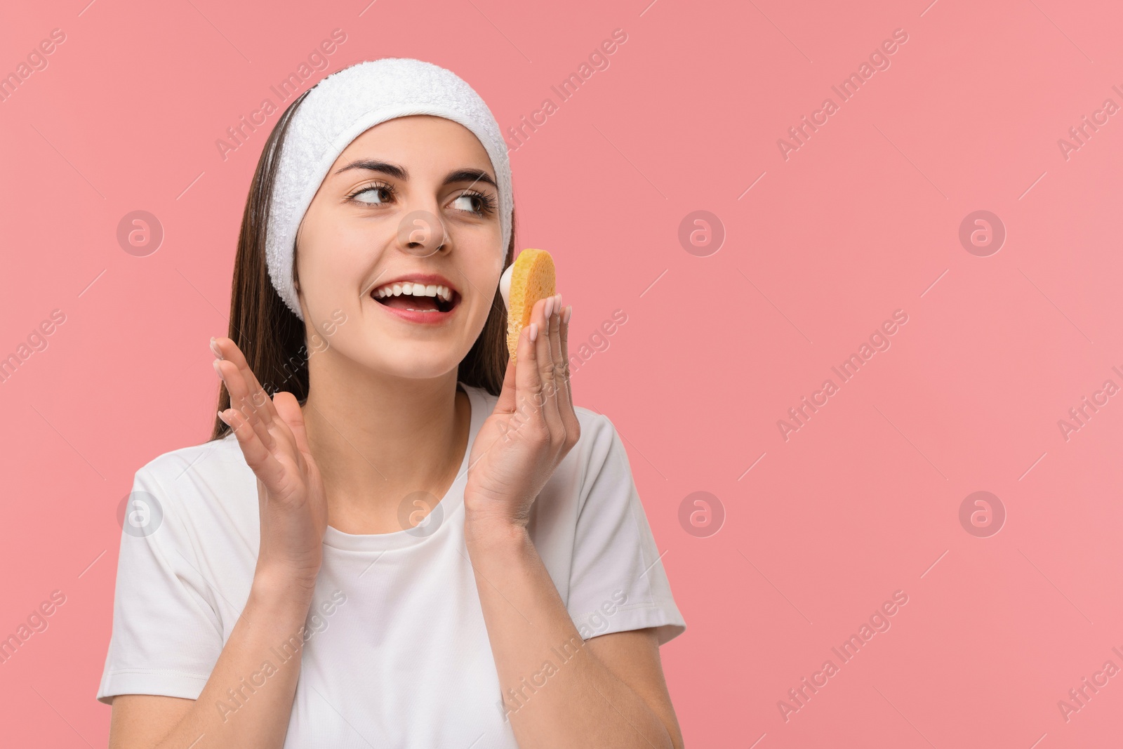 Photo of Young woman with headband washing her face using sponge on pink background, space for text