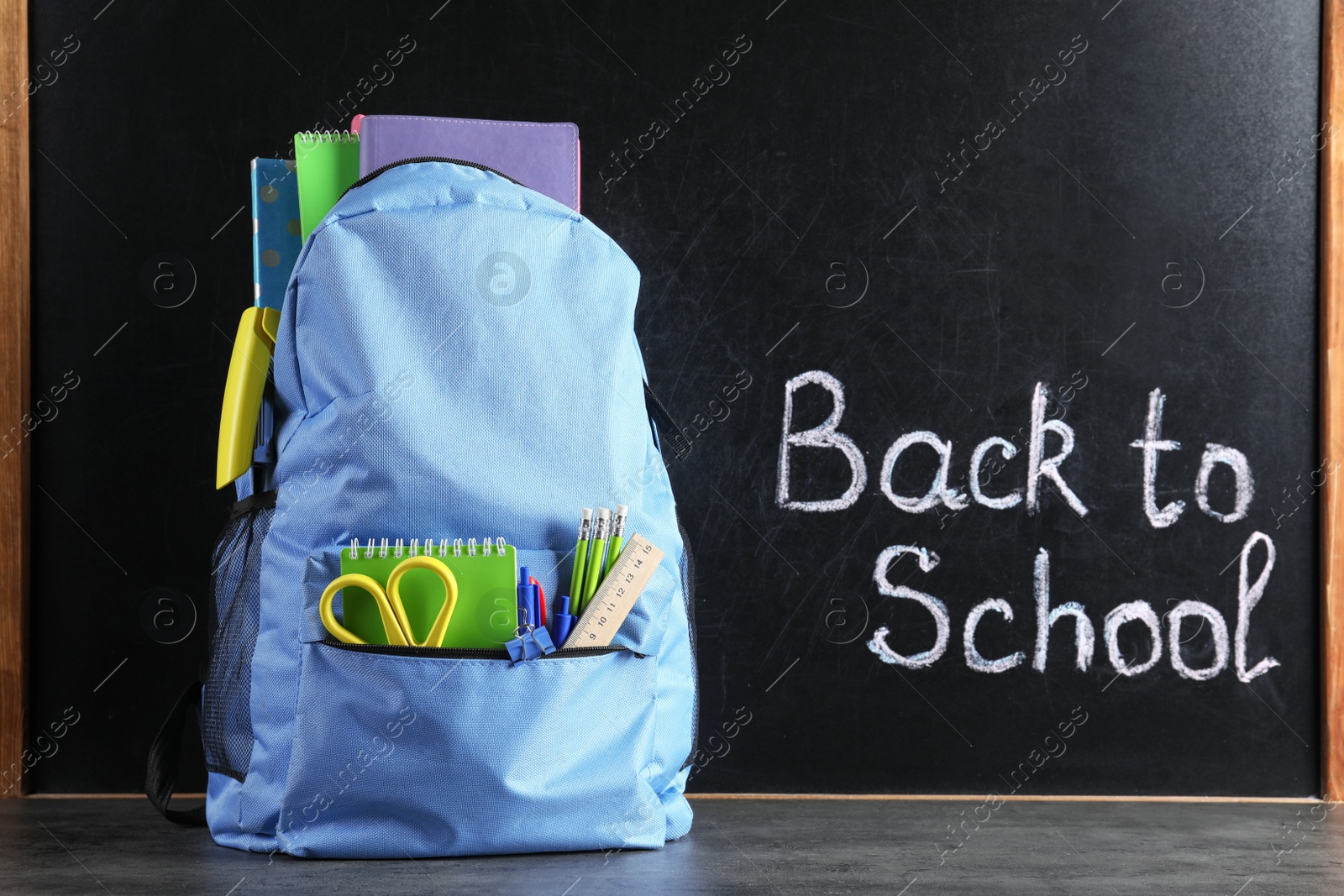 Photo of Backpack with stationery on table against blackboard with written words BACK TO SCHOOL