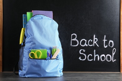 Photo of Backpack with stationery on table against blackboard with written words BACK TO SCHOOL