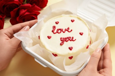 Photo of Woman holding takeaway box with bento cake at beige table, closeup. St. Valentine's day surprise