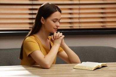 Religious young woman praying over Bible at wooden table indoors