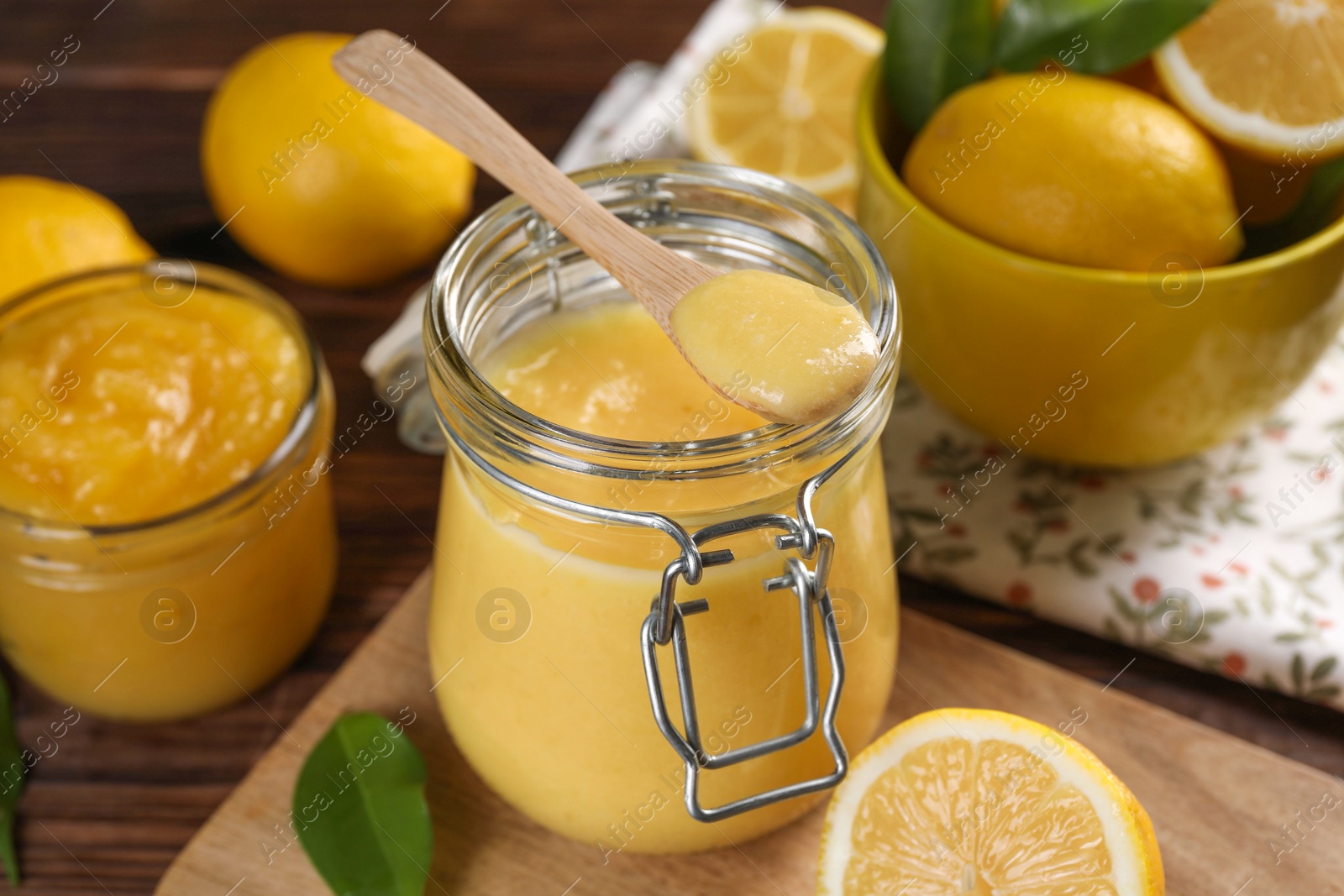 Photo of Delicious lemon curd in glass jars, spoon, fresh citrus fruits and green leaves on wooden table