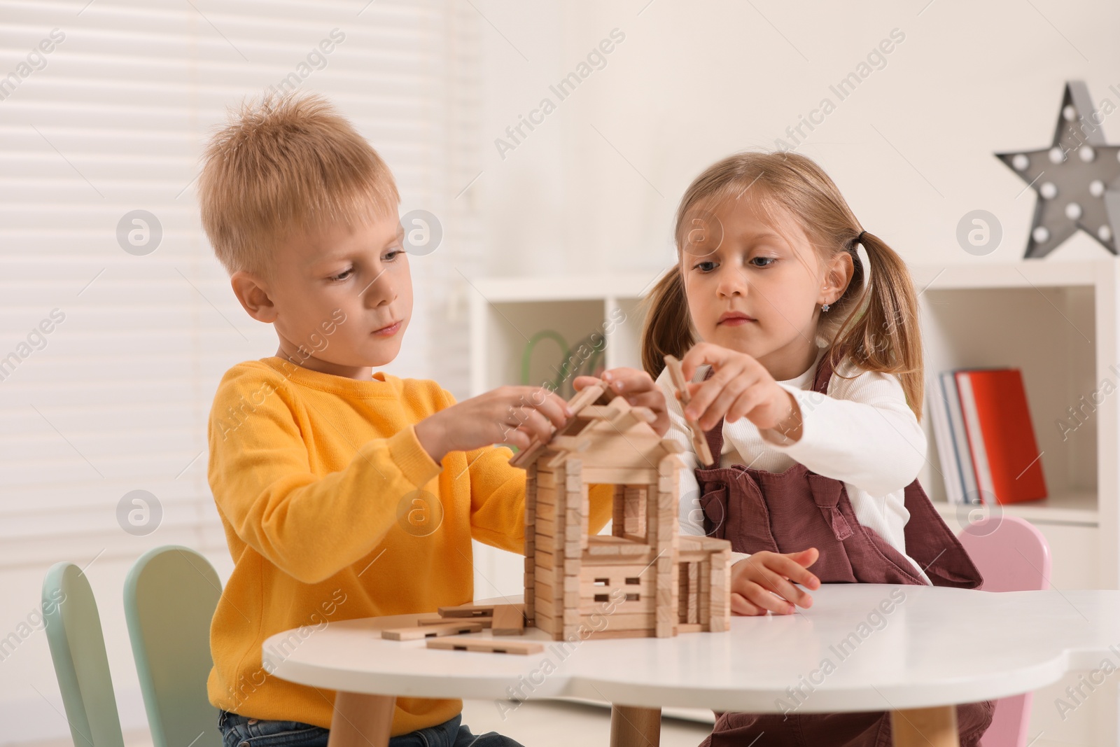 Photo of Little boy and girl playing with wooden house at white table indoors. Children's toys