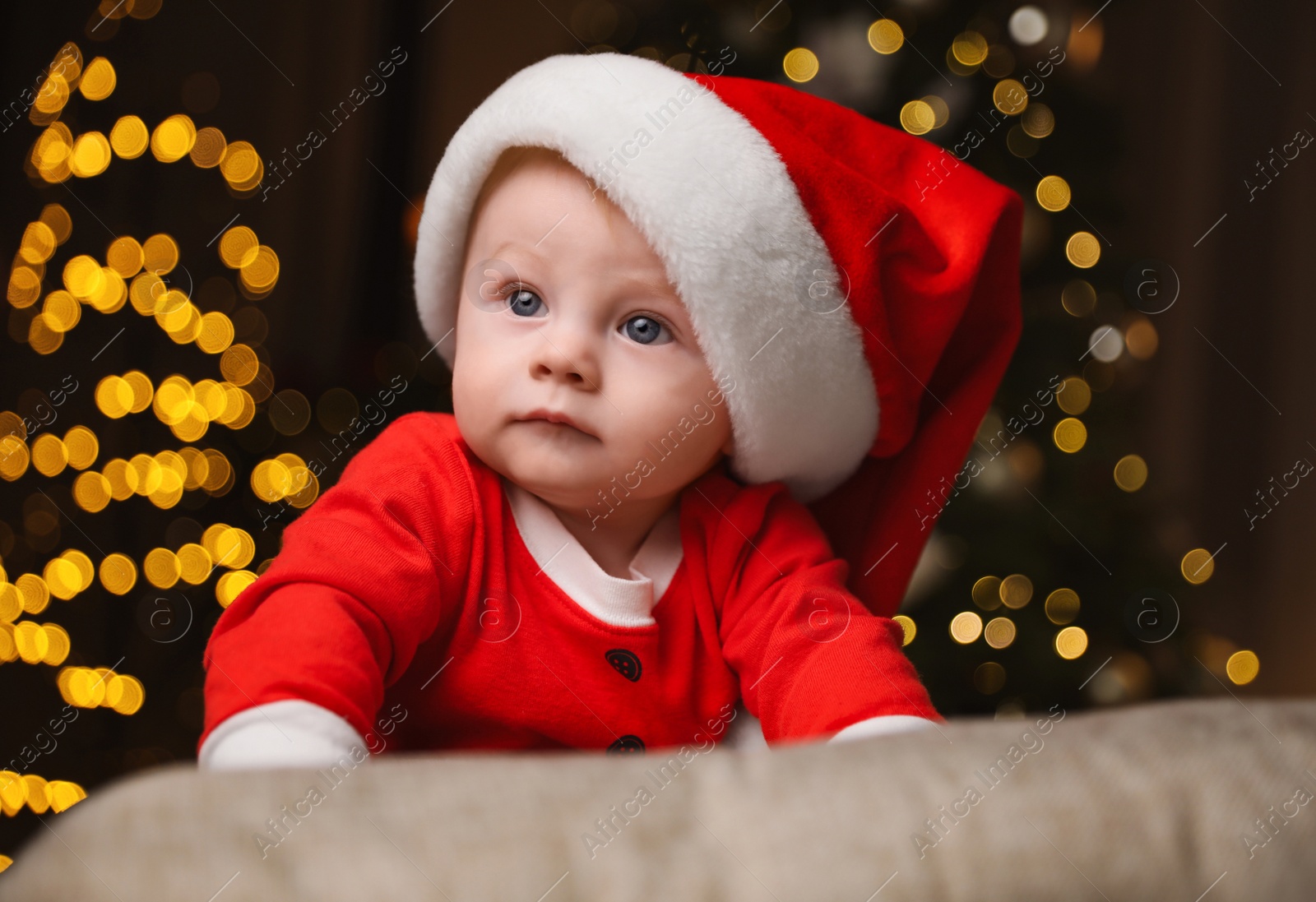 Photo of Cute little baby in Santa Claus suit against blurred festive lights. Christmas celebration