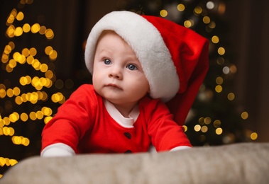 Photo of Cute little baby in Santa Claus suit against blurred festive lights. Christmas celebration