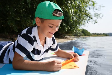 Cute little boy making paper boats on pier near river