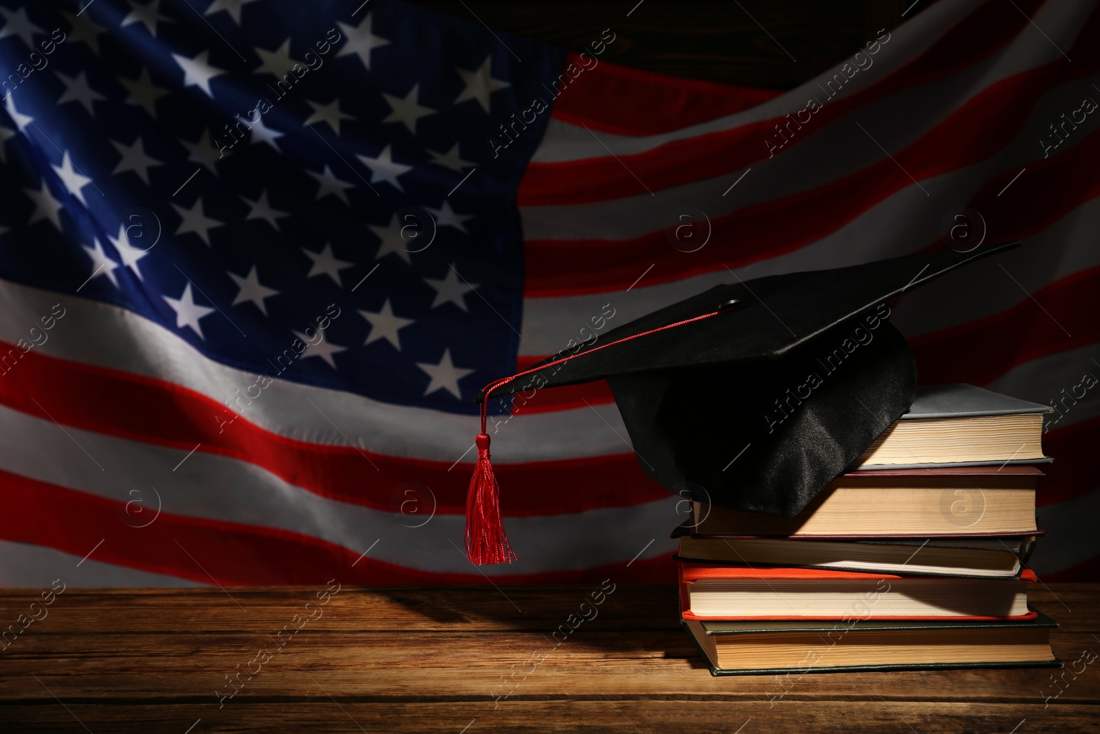 Photo of Graduation hat and books on wooden table against American flag in darkness, space for text