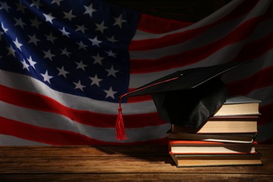 Photo of Graduation hat and books on wooden table against American flag in darkness, space for text