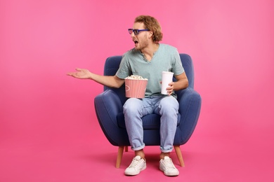 Emotional man with 3D glasses, popcorn and beverage sitting in armchair during cinema show on color background