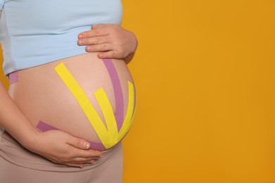 Photo of Pregnant woman with kinesio tapes on her belly against orange background, closeup. Space for text