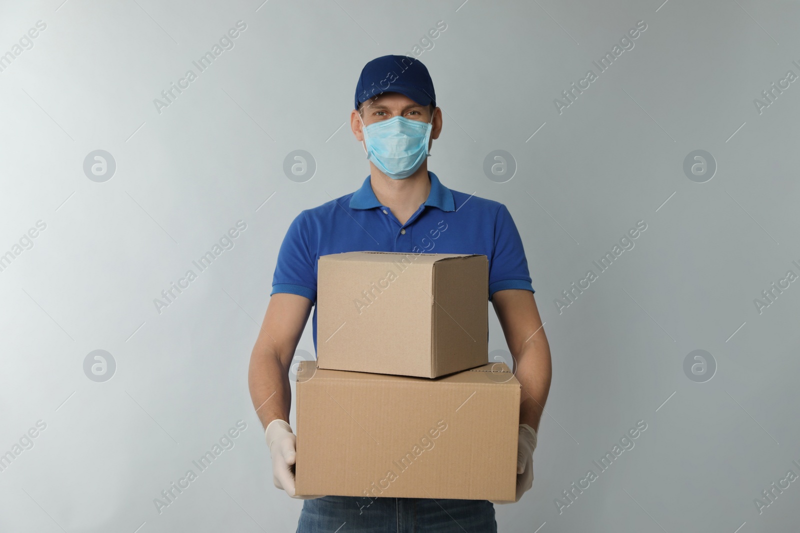 Photo of Courier in protective mask and gloves holding cardboard boxes on light background. Delivery service during coronavirus quarantine