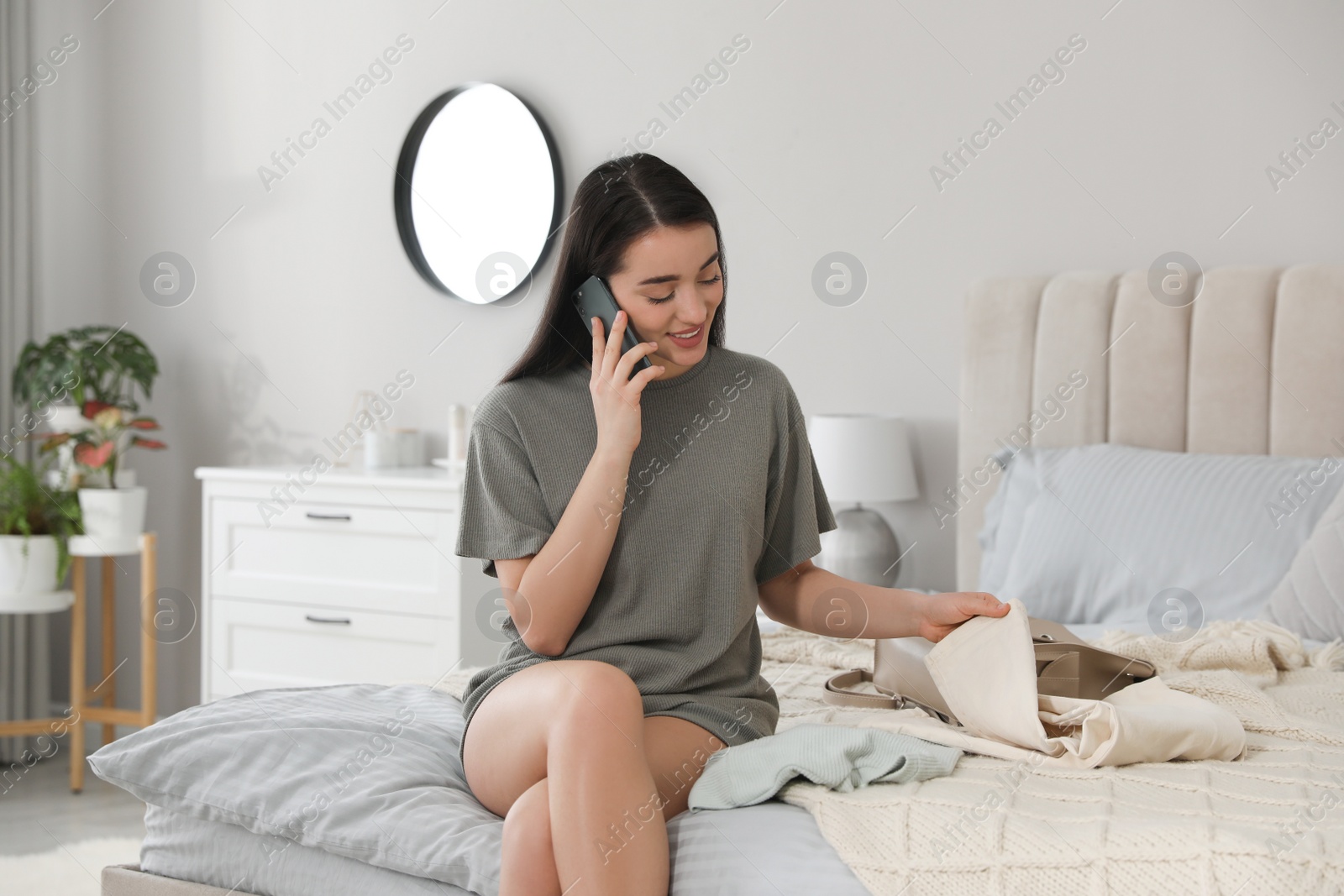 Photo of Young woman talking on phone while choosing clothes for work day at home. Morning routine