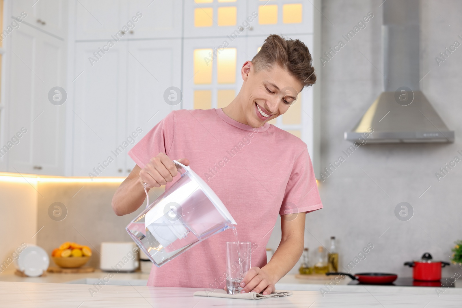 Photo of Happy man pouring water from filter jug into glass at table in kitchen