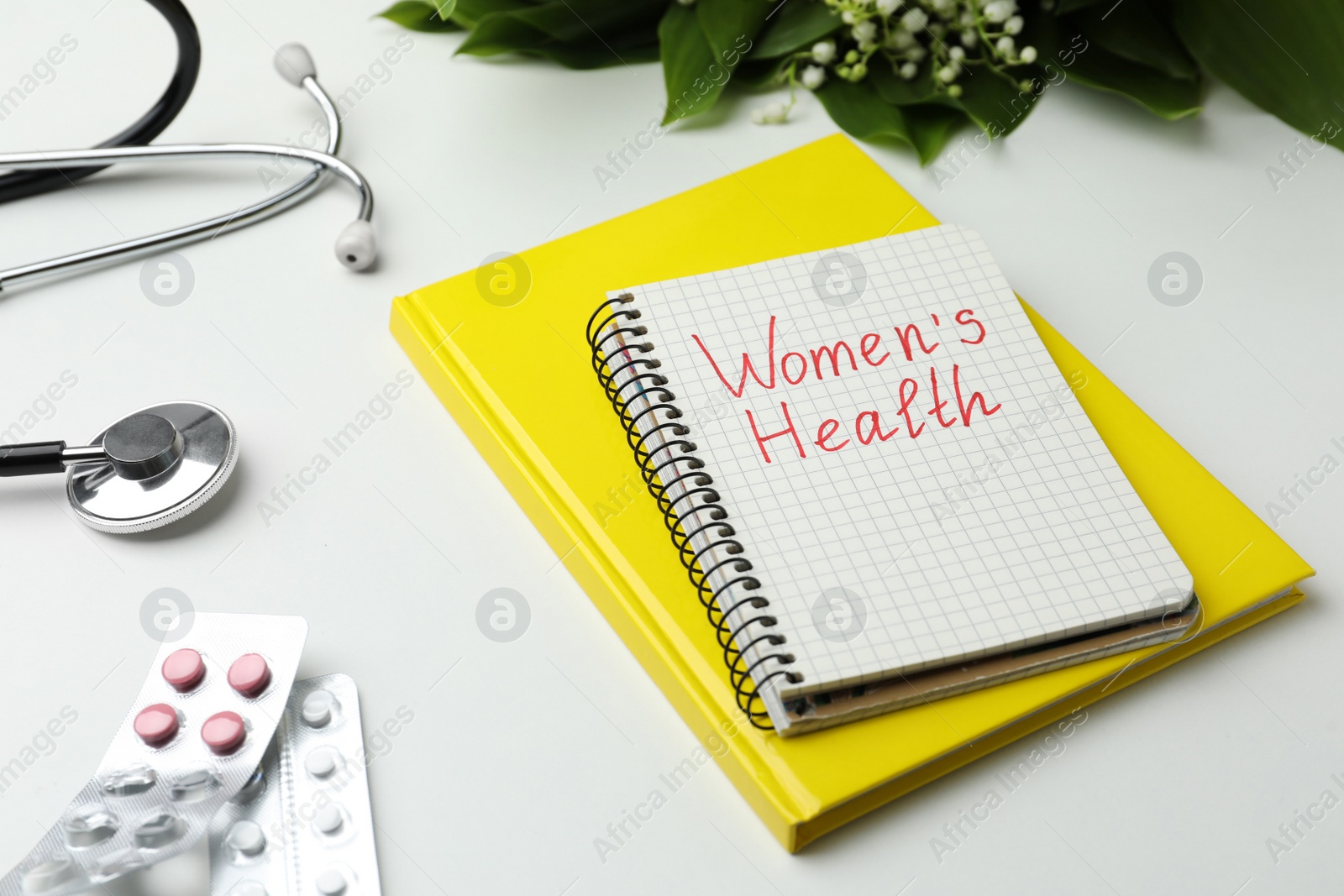 Photo of Notebook with text Women's Health, pills and stethoscope on white table