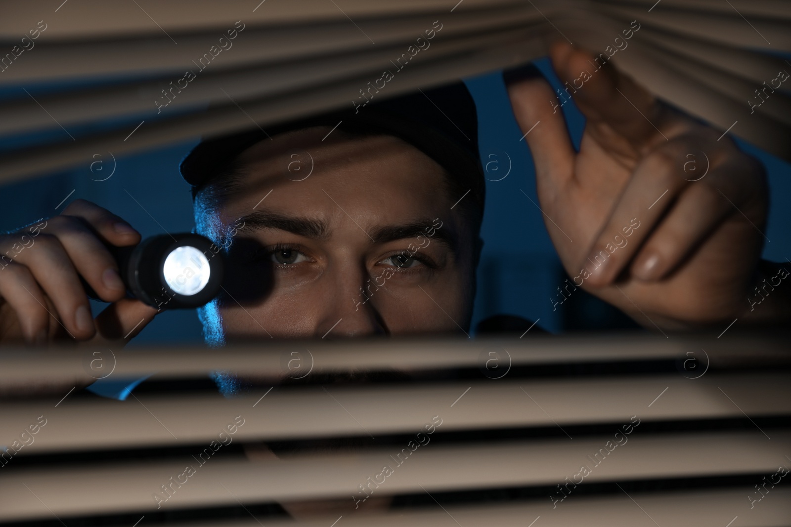 Photo of Male security guard with flashlight looking through window blinds in darkness