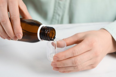 Photo of Woman pouring syrup from bottle into measuring cup at white table, closeup. Cold medicine