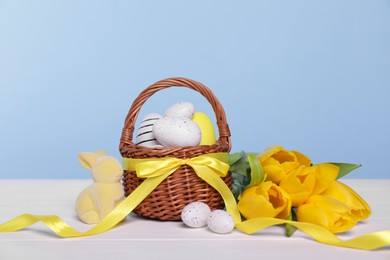 Wicker basket with festively decorated Easter eggs, bunny and beautiful tulips on white wooden table against light blue background
