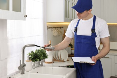 Photo of Young plumber with clipboard examining faucet in kitchen