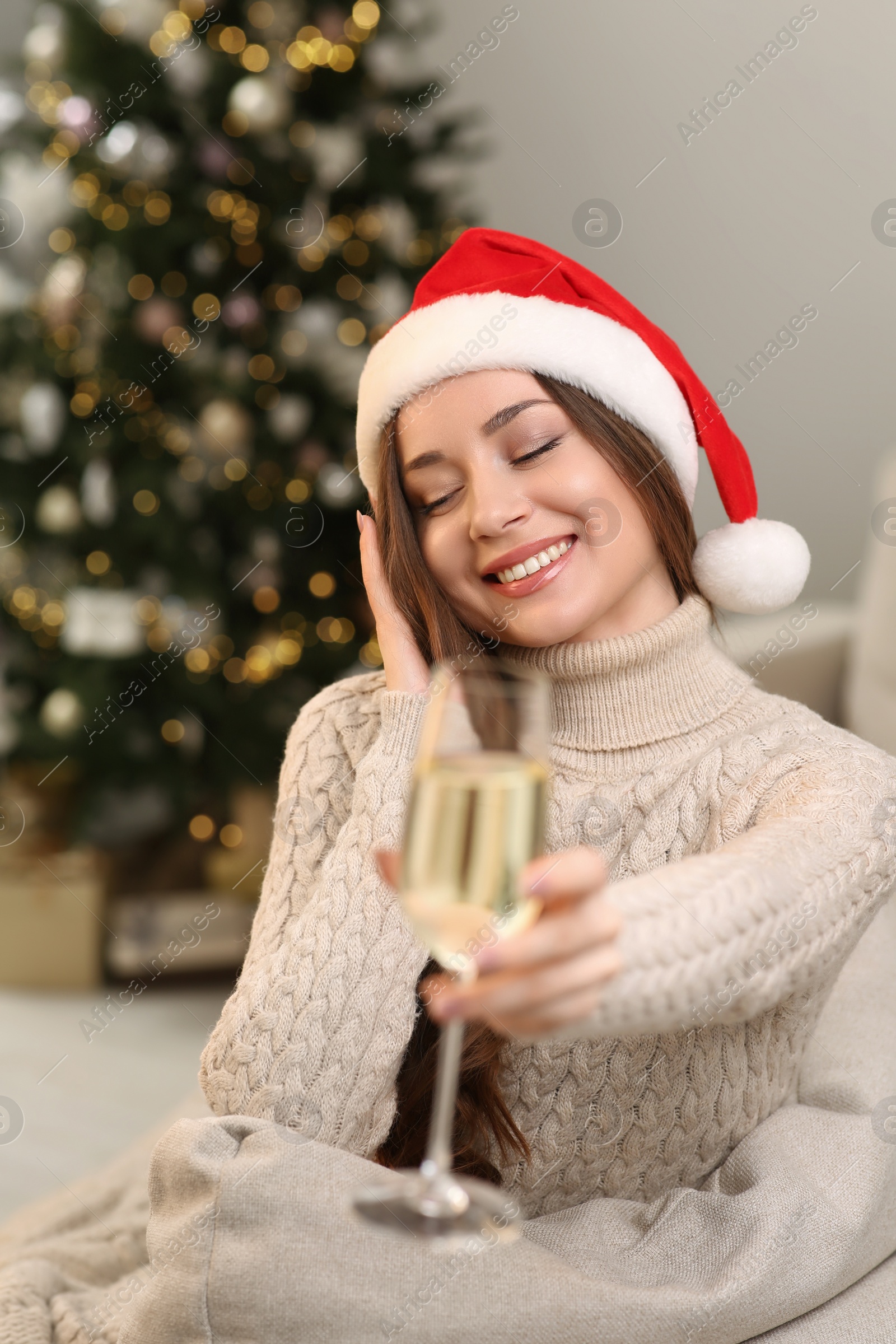 Photo of Beautiful young smiling woman in Santa hat with glass of sparkling wine at home