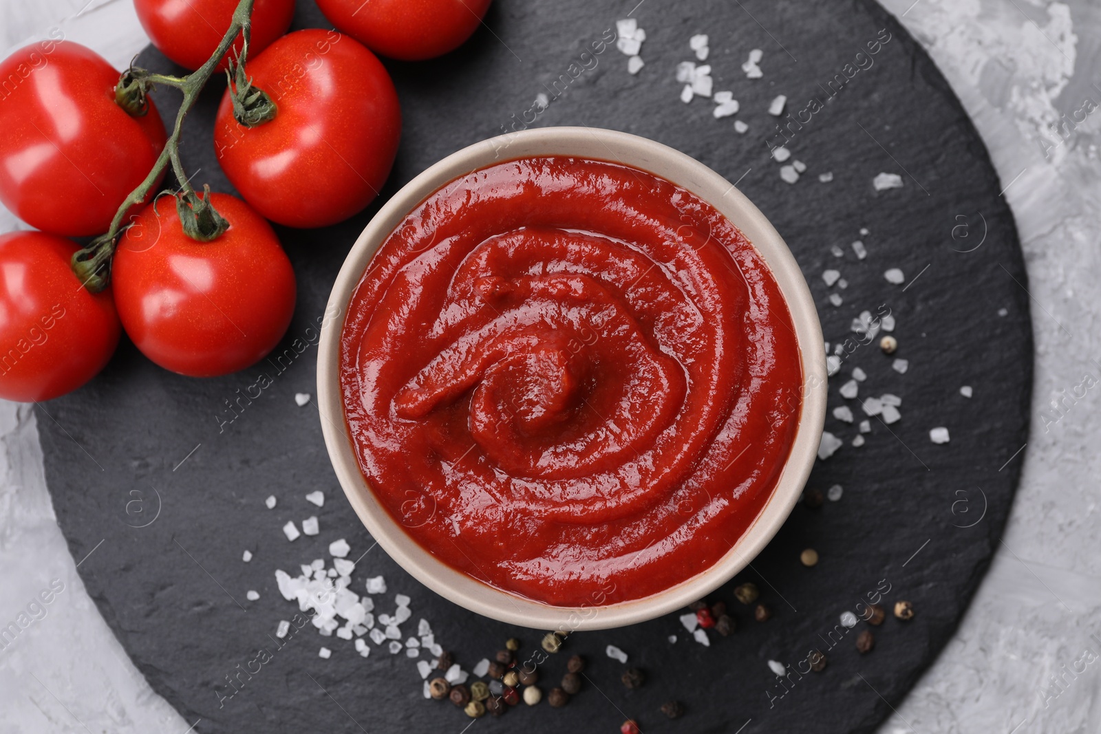 Photo of Organic ketchup in bowl, fresh tomatoes and spices on grey textured table, top view. Tomato sauce