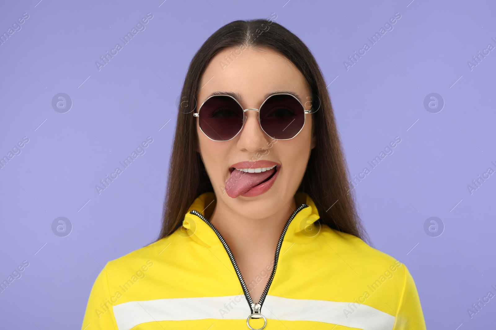 Photo of Happy woman in sunglasses showing her tongue on violet background